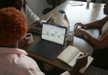 Three people in a meeting at a table discussing schedule on their Microsoft laptop